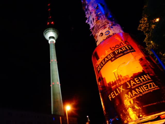 Plakat auf Litfaßsäule mit Fernsehturm im Hintergrund bei Nacht